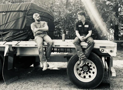 Two people sit on a truck bed with forest trees in background during daytime