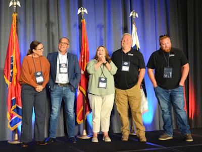 Five professionals wearing badges stand together on stage in front of flags at a conference event.