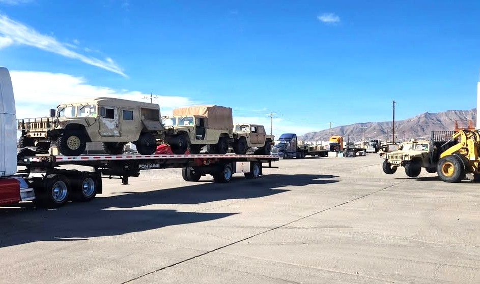 Flatbed trailer transporting tan military vehicles at an industrial facility with mountains in background