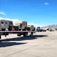Flatbed trailer transporting tan military vehicles at an industrial facility with mountains in background