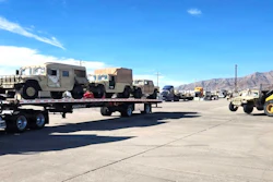 Flatbed trailer transporting tan military vehicles at an industrial facility with mountains in background