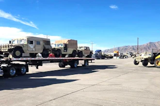 Flatbed trailer transporting tan military vehicles at an industrial facility with mountains in background