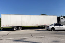 Sheriff's patrol car positioned alongside a large white commercial semi-truck on an open road under clear blue sky