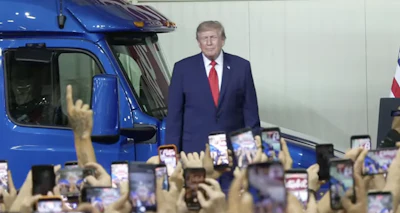 Man in navy suit standing beside blue truck at public event with crowd holding phones