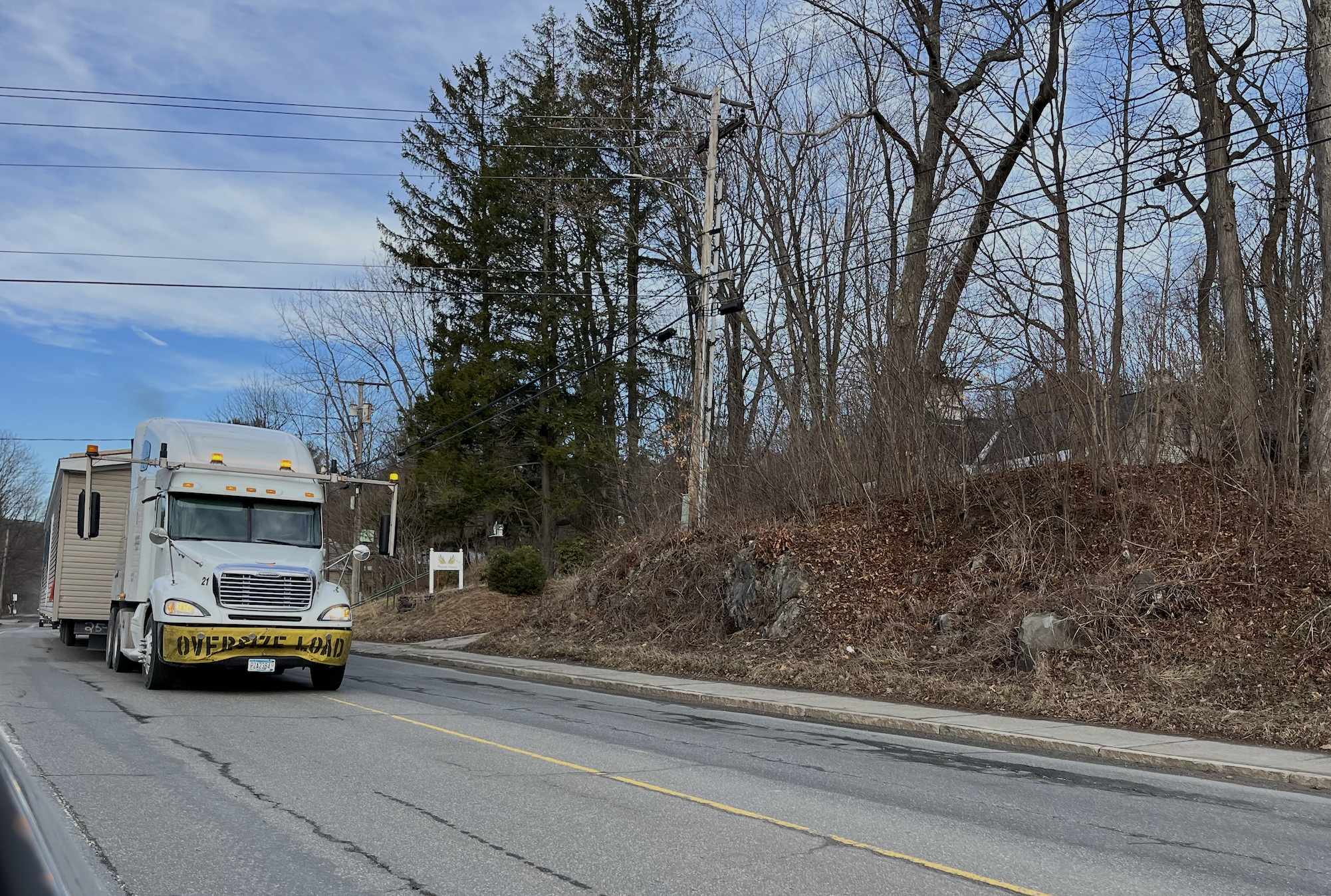 White moving truck with yellow trim parked on residential road lined with bare trees and hillside