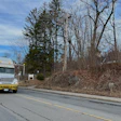 White moving truck with yellow trim parked on residential road lined with bare trees and hillside