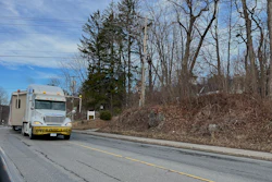 White moving truck with yellow trim parked on residential road lined with bare trees and hillside
