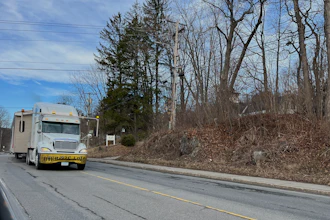 White moving truck with yellow trim parked on residential road lined with bare trees and hillside