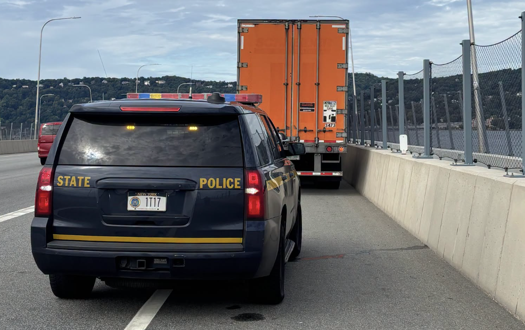 State Police vehicle escorting orange commercial truck on highway with concrete barriers and bridge visible