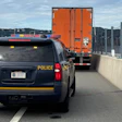 State Police vehicle escorting orange commercial truck on highway with concrete barriers and bridge visible