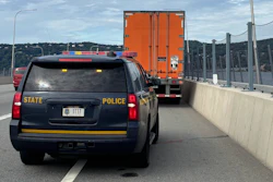 State Police vehicle escorting orange commercial truck on highway with concrete barriers and bridge visible