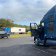 Blue semi-truck with open driver door parked in yard beside pickup truck and trailers
