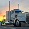 Silver Peterbilt semi-truck with dual exhaust stacks parked at sunset with American flag visible