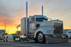 Silver Peterbilt semi-truck with dual exhaust stacks parked at sunset with American flag visible