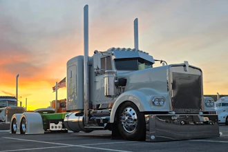 Silver Peterbilt semi-truck with dual exhaust stacks parked at sunset with American flag visible