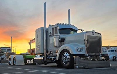 Silver Peterbilt semi-truck with dual exhaust stacks parked at sunset with American flag visible