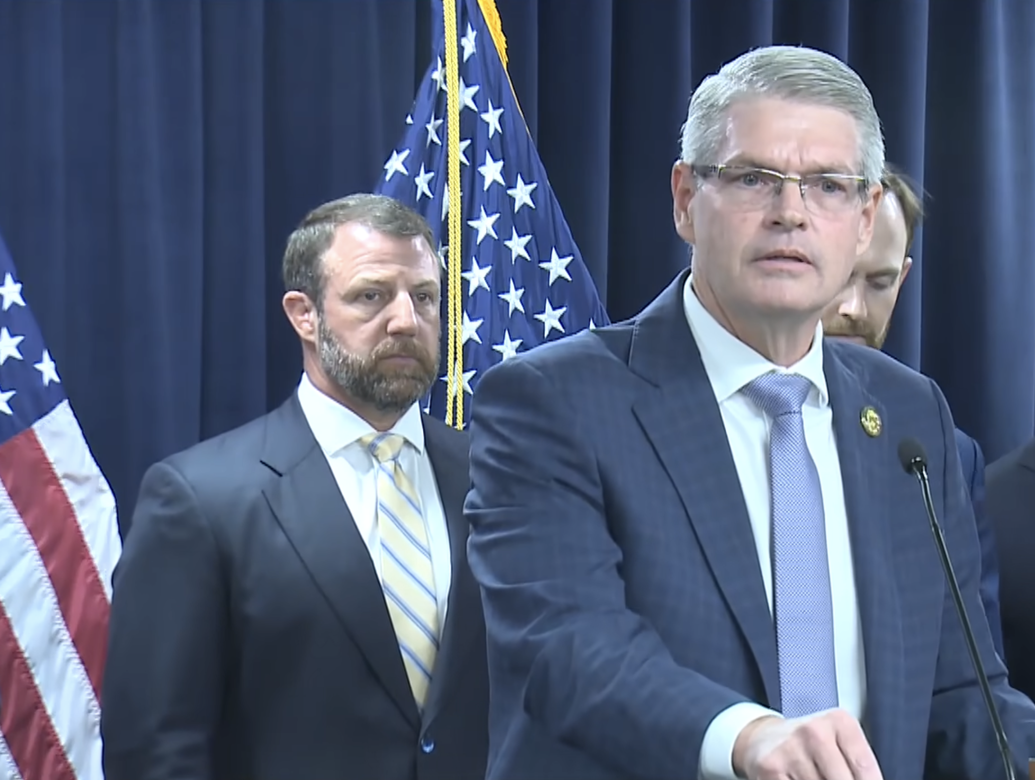 Three officials at a podium during a formal press conference with American flags in the background.