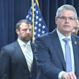 Three officials at a podium during a formal press conference with American flags in the background.