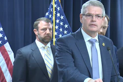 Three officials at a podium during a formal press conference with American flags in the background.