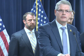Three officials at a podium during a formal press conference with American flags in the background.