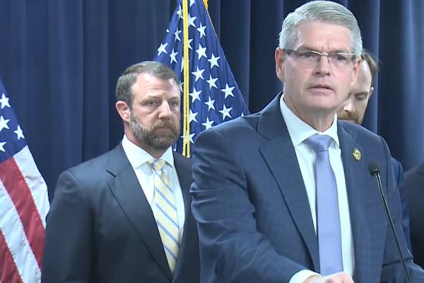 Three officials at a podium during a formal press conference with American flags in the background.