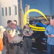 Law enforcement officers and civilians gathered near a yellow and blue striped bus during an outdoor community event.