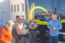 Law enforcement officers and civilians gathered near a yellow and blue striped bus during an outdoor community event.