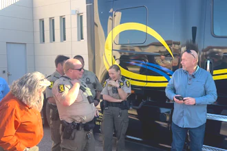 Law enforcement officers and civilians gathered near a yellow and blue striped bus during an outdoor community event.