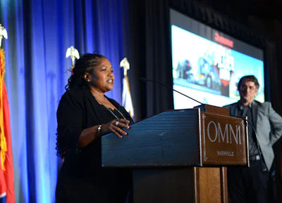 Woman speaking at OMNI Nashville podium during conference presentation with screen display in background