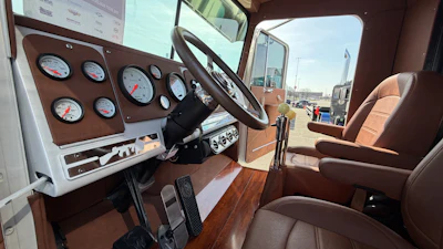 Classic truck cab interior with steering wheel, analog gauges, brown seats, and industrial port view through windshield