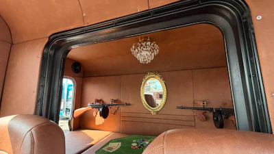 Luxury vehicle interior with tan leather seating, crystal chandelier, ornate mirror, and decorative firearms on paneled walls