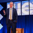 Professional speaker in business suit presenting on stage with blue geometric backdrop design