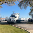 Fleet of white commercial semi-trucks parked under large trees in a logistics yard on a clear day
