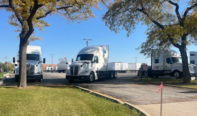 Fleet of white commercial semi-trucks parked under large trees in a logistics yard on a clear day