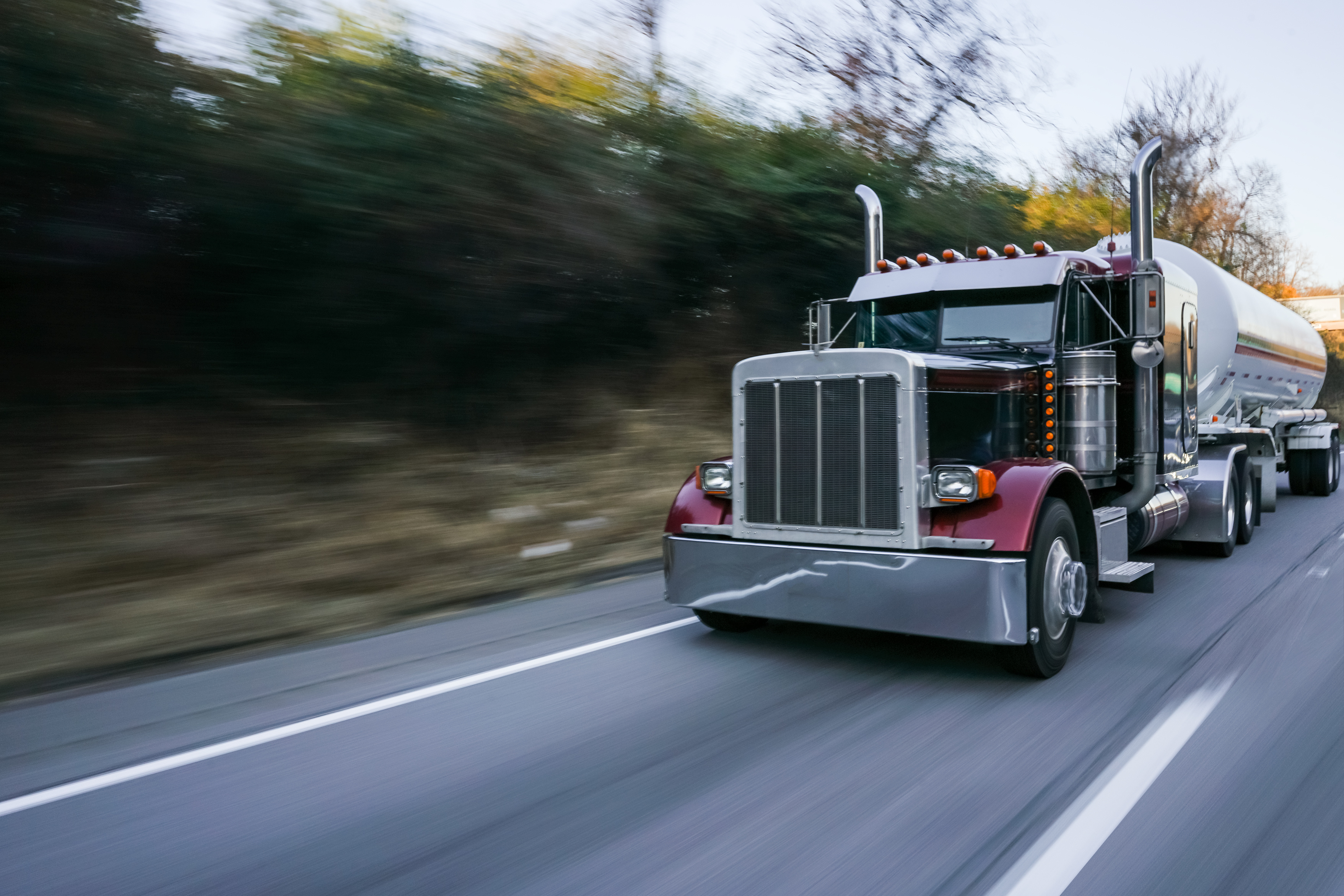 Red and silver semi-truck with tanker trailer driving on highway with blurred trees in background