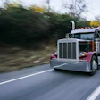 Red and silver semi-truck with tanker trailer driving on highway with blurred trees in background