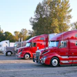 Fleet of red and white semi-trucks parked in outdoor lot with autumn trees in background