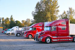 Fleet of red and white semi-trucks parked in outdoor lot with autumn trees in background