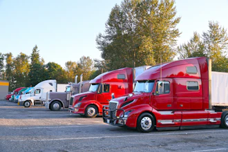 Fleet of red and white semi-trucks parked in outdoor lot with autumn trees in background