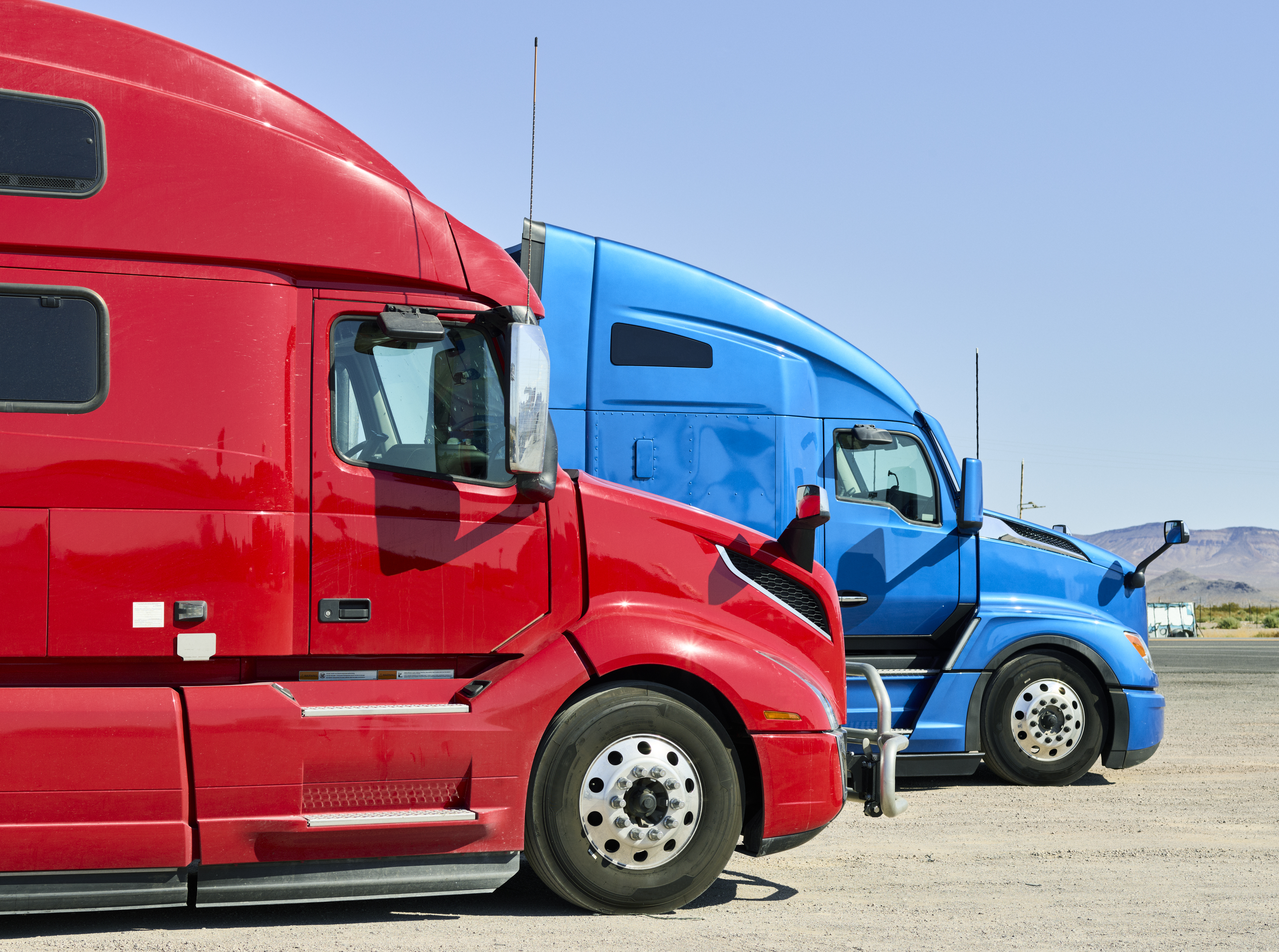 Red and blue semi-trucks parked side by side in an outdoor lot with clear sky