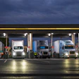 Multiple commercial semi-trucks parked under a covered loading facility at dusk with illuminated signs