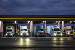 Multiple commercial semi-trucks parked under a covered loading facility at dusk with illuminated signs