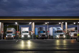 Multiple commercial semi-trucks parked under a covered loading facility at dusk with illuminated signs